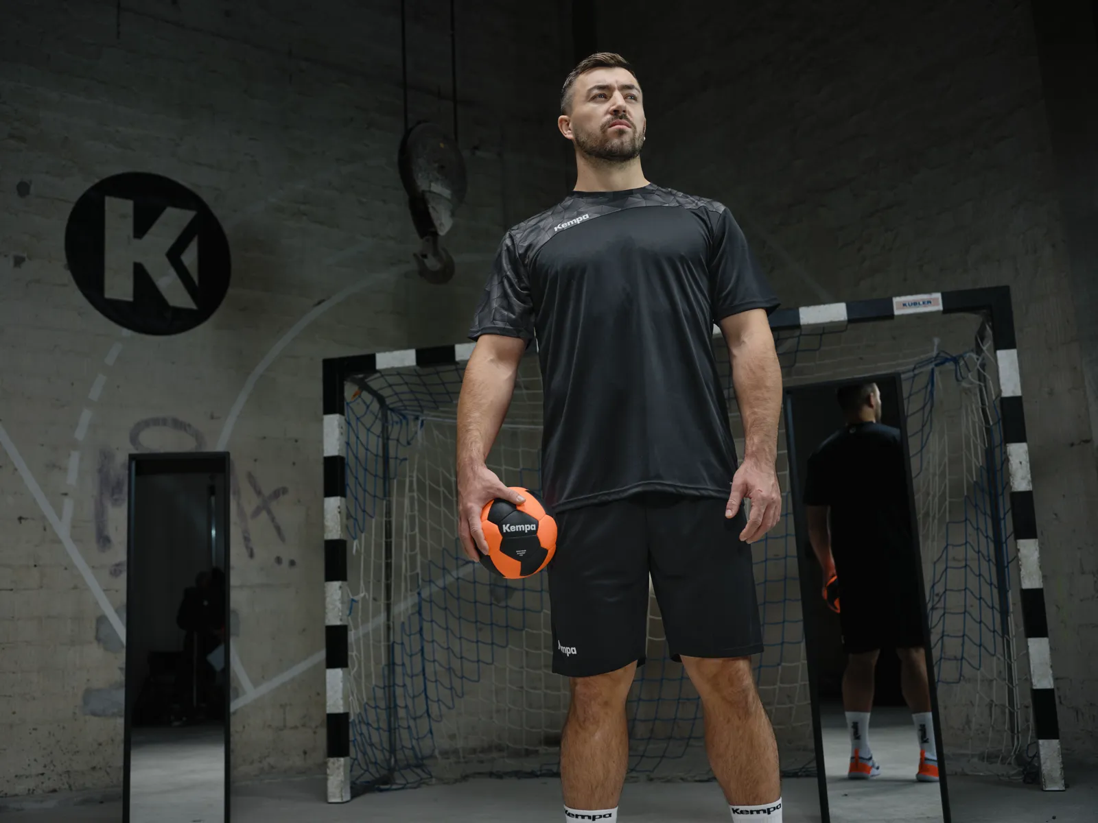 Handballprofi mit orangem Ball in der Industriehalle, Kampagne 2026